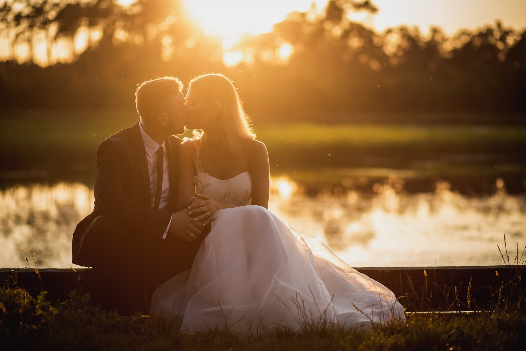 Para młoda w ślubnych strojach całuje się siedząc na drewnianym pomoście nad wodą, w tle zachodzące słońce tworzy złocistą poświatę.