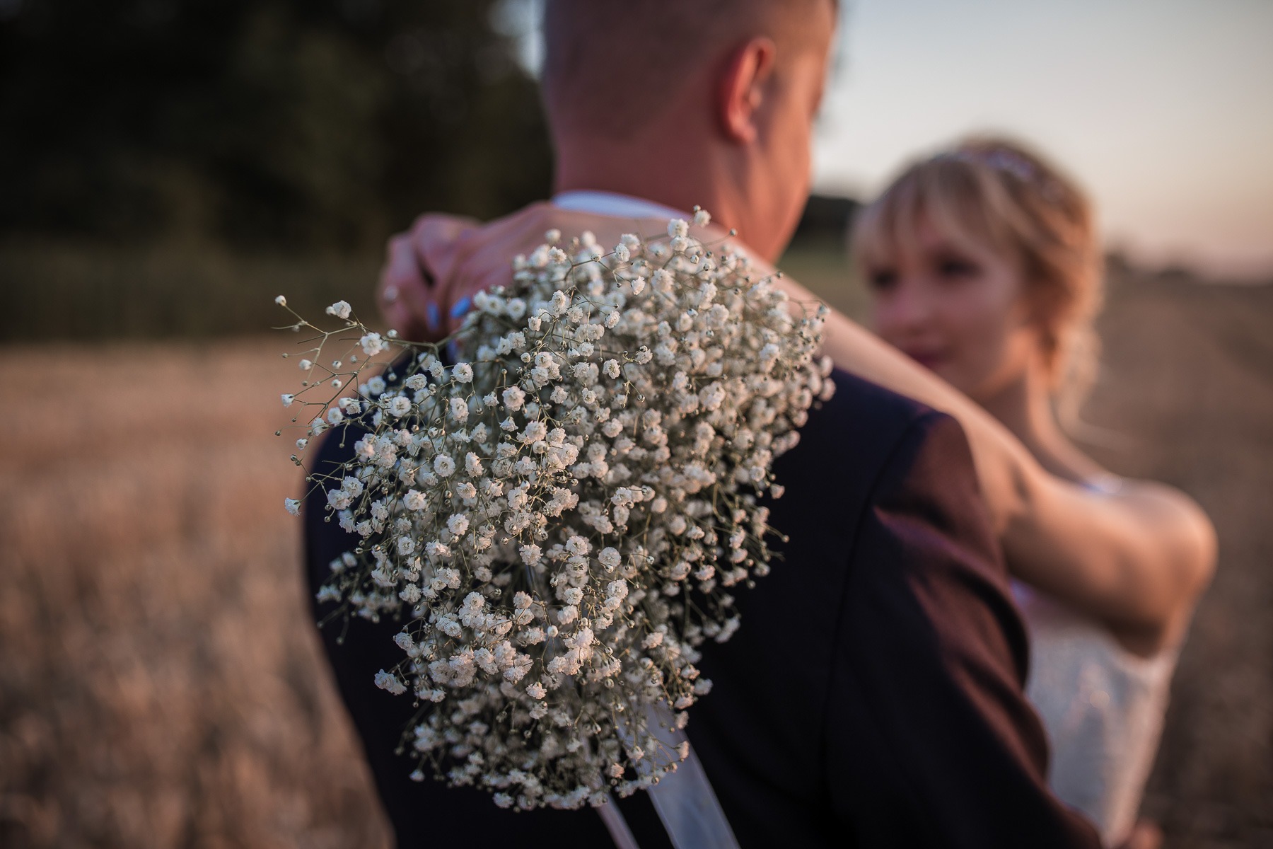 Para młoda w objęciach w plenerze, bukiet gipsówki w dłoniach, delikatne światło zachodzącego słońca.