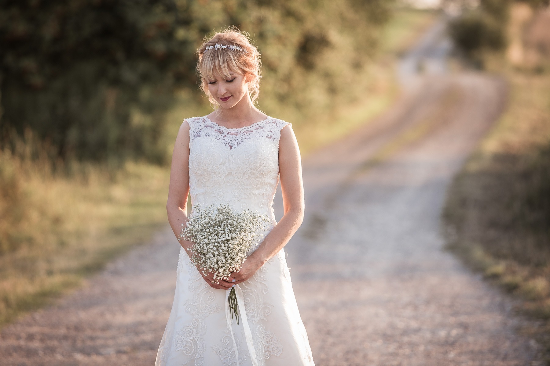 Panna młoda w koronkowej sukni ślubnej, trzymająca bukiet gipsówki, na polnej drodze w ciepłym, naturalnym świetle.