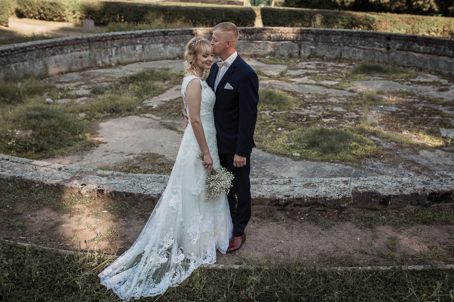 Panna młoda w koronkowej sukni ślubnej i pan młody w granatowym garniturze stoją na tle kamiennego murku, pan młody całuje pannę młodą w czoło.