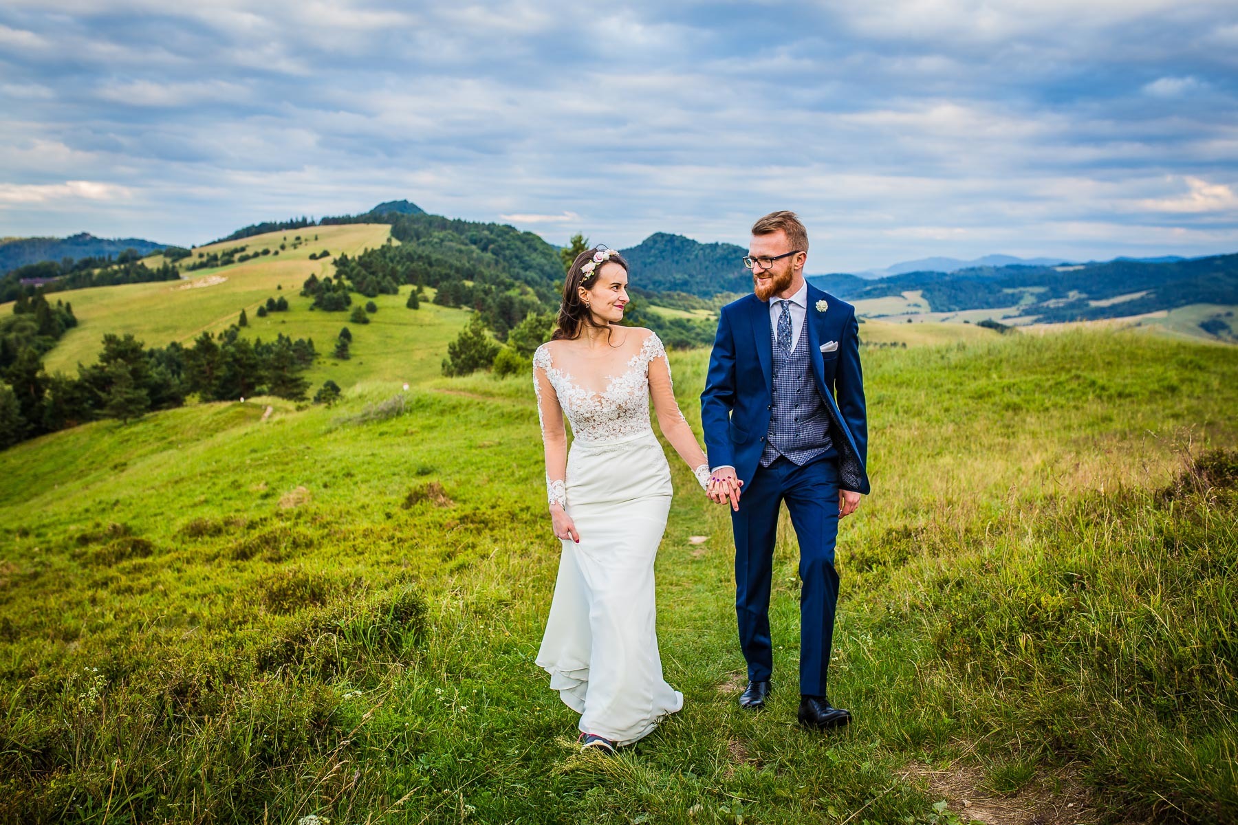 Para młoda, on w granatowym garniturze i ona w białej sukni z koronkową górą, spaceruje po zielonym wzgórzu z widokiem na góry i pochmurne niebo.