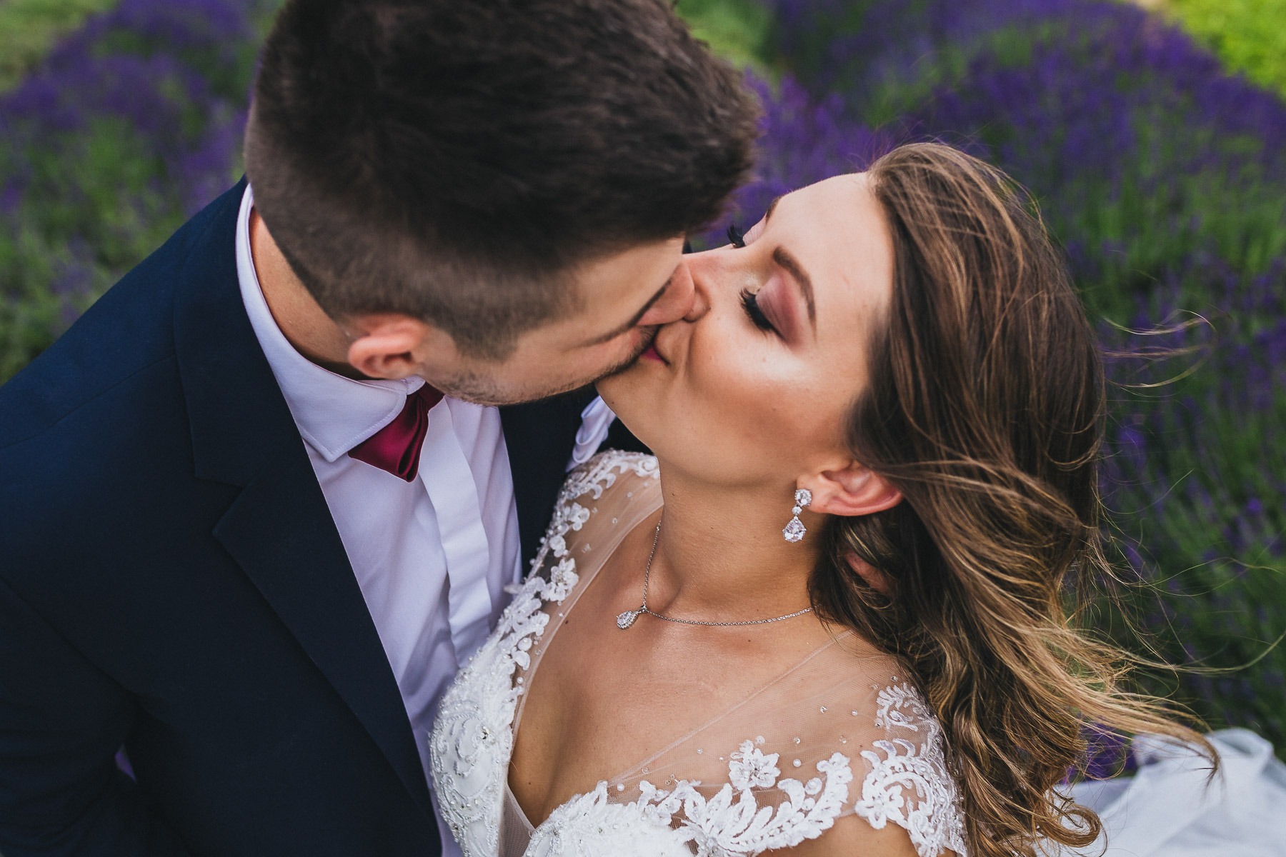 Pocałunek nowożeńców na tle kwitnącej lawendy, panna młoda w sukni z koronkowymi aplikacjami, widoczne kolczyki i naszyjnik z zawieszką.