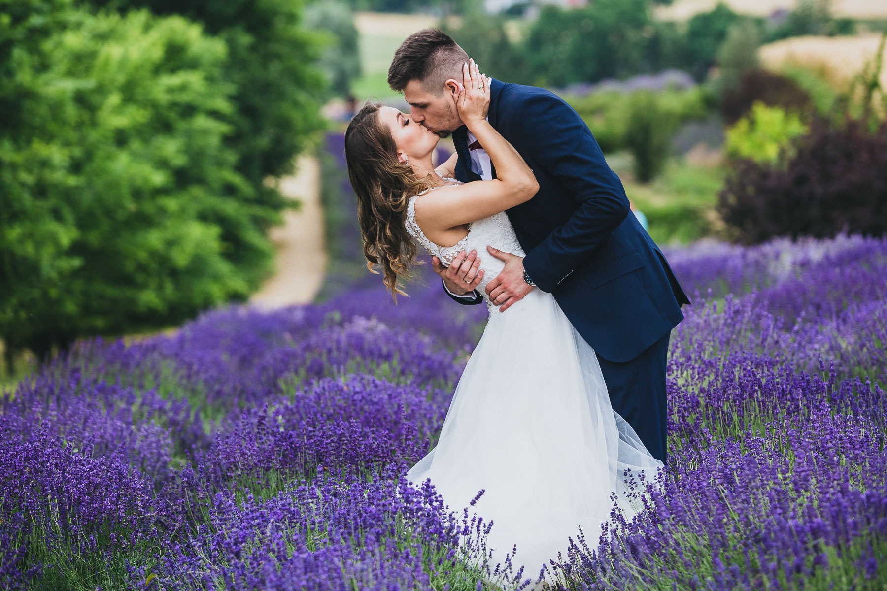 Pocałunek młodej pary w polu kwitnącej lawendy, panna młoda w białej sukni z koronkową górą, pan młody w granatowym garniturze.