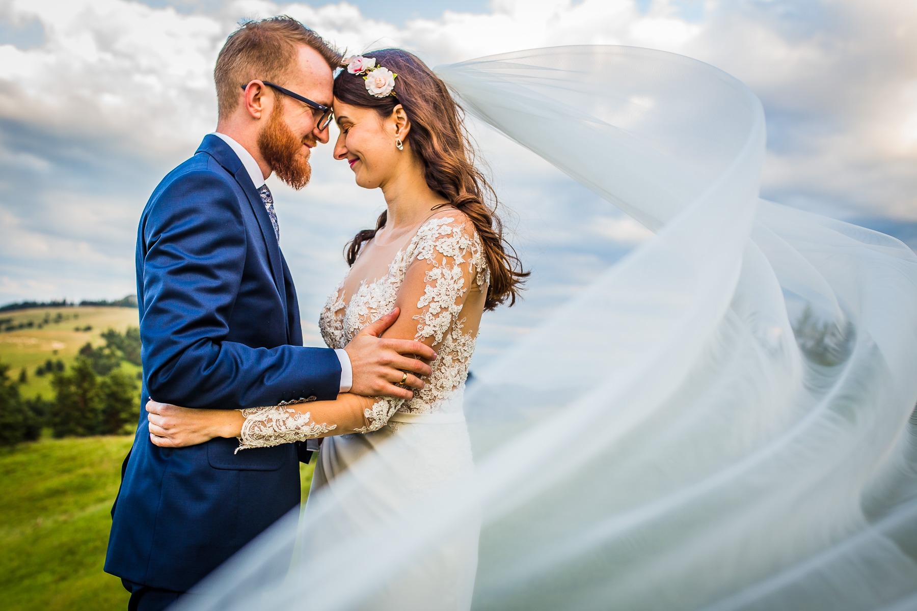 Uśmiechnięta para młoda, on w granatowym garniturze i okularach, ona w koronkowej sukni ślubnej i wianku, obejmuje się na tle zielonych wzgórz i pochmurnego nieba, z powiewającym welonem.