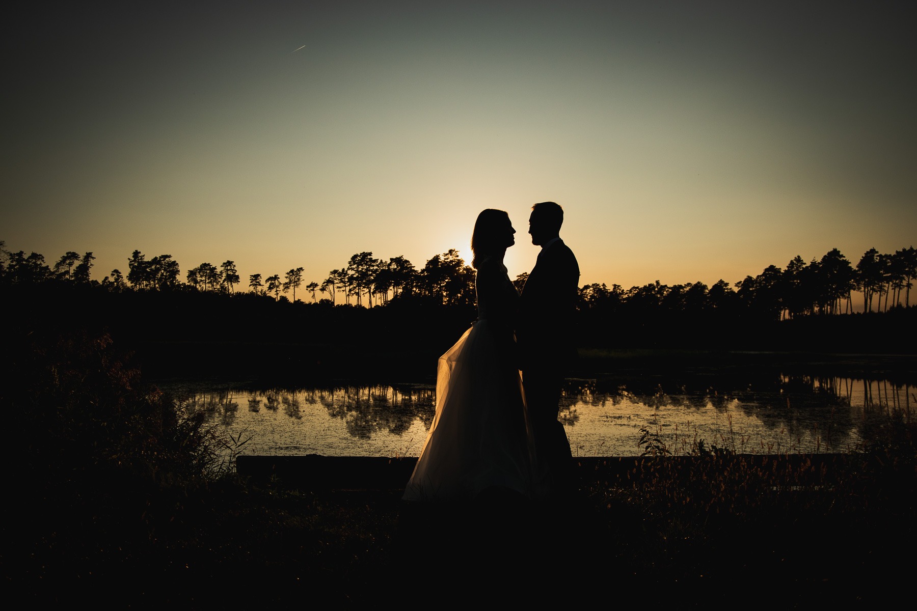 Sylwetki pary młodej na tle zachodzącego słońca odbijającego się w tafli jeziora, otoczonego drzewami.