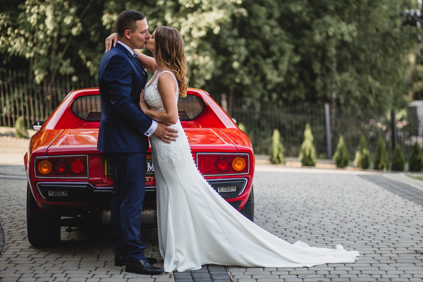 Pocałunek pary młodej na tle czerwonego, sportowego samochodu Ferrari Dino 308 GT4 z długim trenem sukni ślubnej na brukowanym podjeździe.