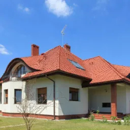 Dom jednorodzinny z czerwoną dachówką ceramiczną, balkonem z metalową balustradą i białymi ścianami wykończonymi ceglaną podmurówką w słoneczny dzień.