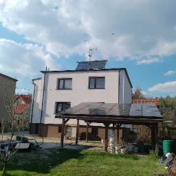 Dwukondygnacyjny dom z białymi ścianami, brązowym cokołem, kolektorami słonecznymi na dachu i zadaszeniem z paneli fotowoltaicznych w ogrodzie, widok w słoneczny dzień.