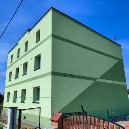 Trzypiętrowy budynek z ocieploną elewacją w odcieniach zieleni, z geometrycznymi wzorami, widoczny z perspektywy ulicy, z ogrodzeniem w pierwszym planie.