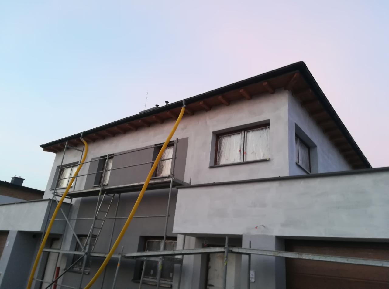 Dwupiętrowy dom w trakcie prac elewacyjnych, z widocznymi rusztowaniami, zabezpieczonymi oknami folią i fragmentami pomalowanymi na szaro, z żółtą rurą spustową przy rynnie.