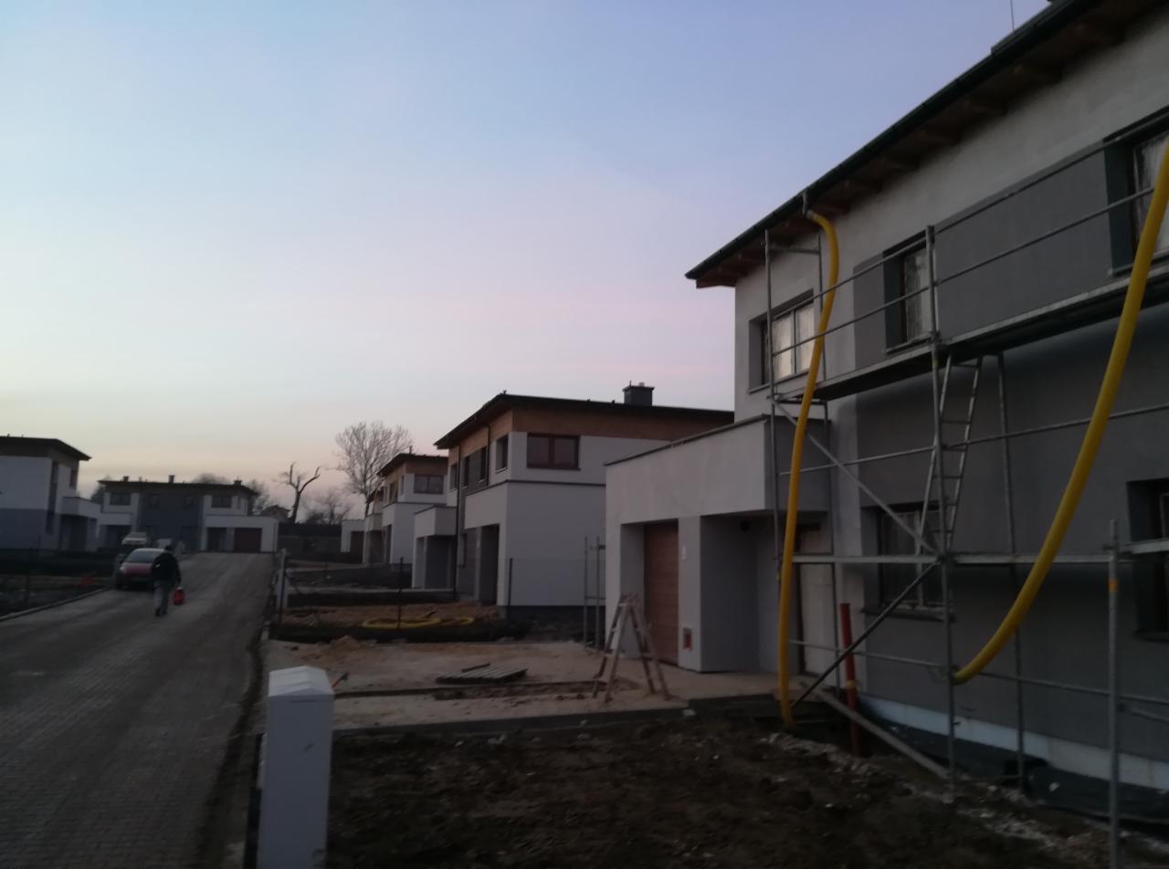 Szeregówka domów jednorodzinnych w trakcie prac elewacyjnych; widoczne rusztowania i żółty wąż wentylacyjny, fragmenty elewacji w szarym podkładzie, w tle ulica i osoba z wiadrem.