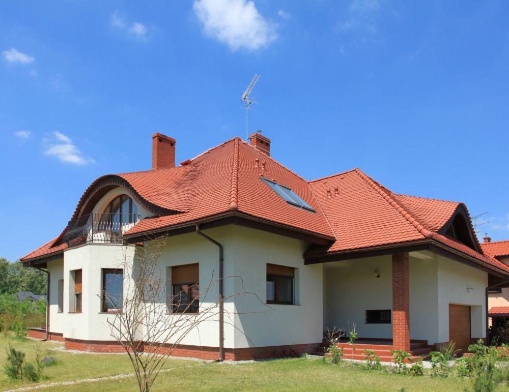 Dom jednorodzinny z czerwoną dachówką ceramiczną, balkonem z metalową balustradą i białymi ścianami wykończonymi ceglaną podmurówką w słoneczny dzień.