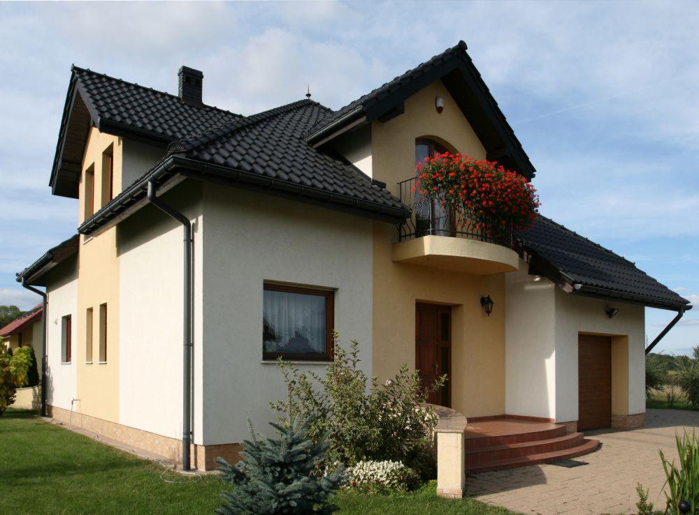 Dwukondygnacyjny dom jednorodzinny z kremowo-żółtą elewacją, czarnym dachem z rynnami i balkonem ozdobionym czerwonymi kwiatami, widok z poziomu trawnika.