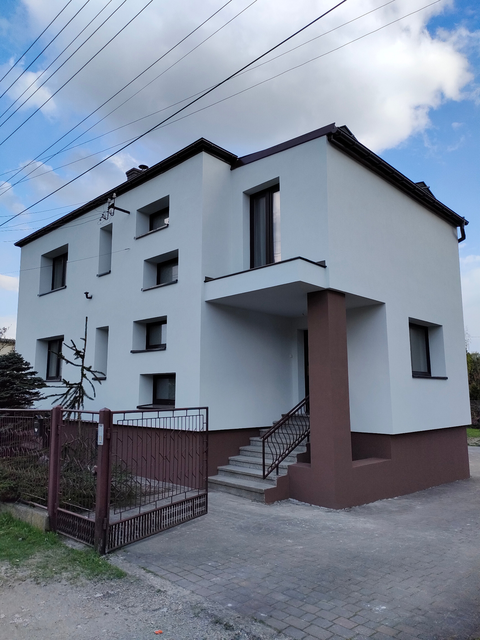 Dwukondygnacyjny dom z białą elewacją i brązowymi akcentami, widok z poziomu gruntu na fasadę frontową, z otwartą bramą wjazdową i schodami wejściowymi.