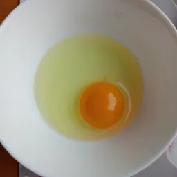 Surowe jajo z intensywnie pomarańczowym żółtkiem w białej miseczce na podkładce z motywem kwiatowym.