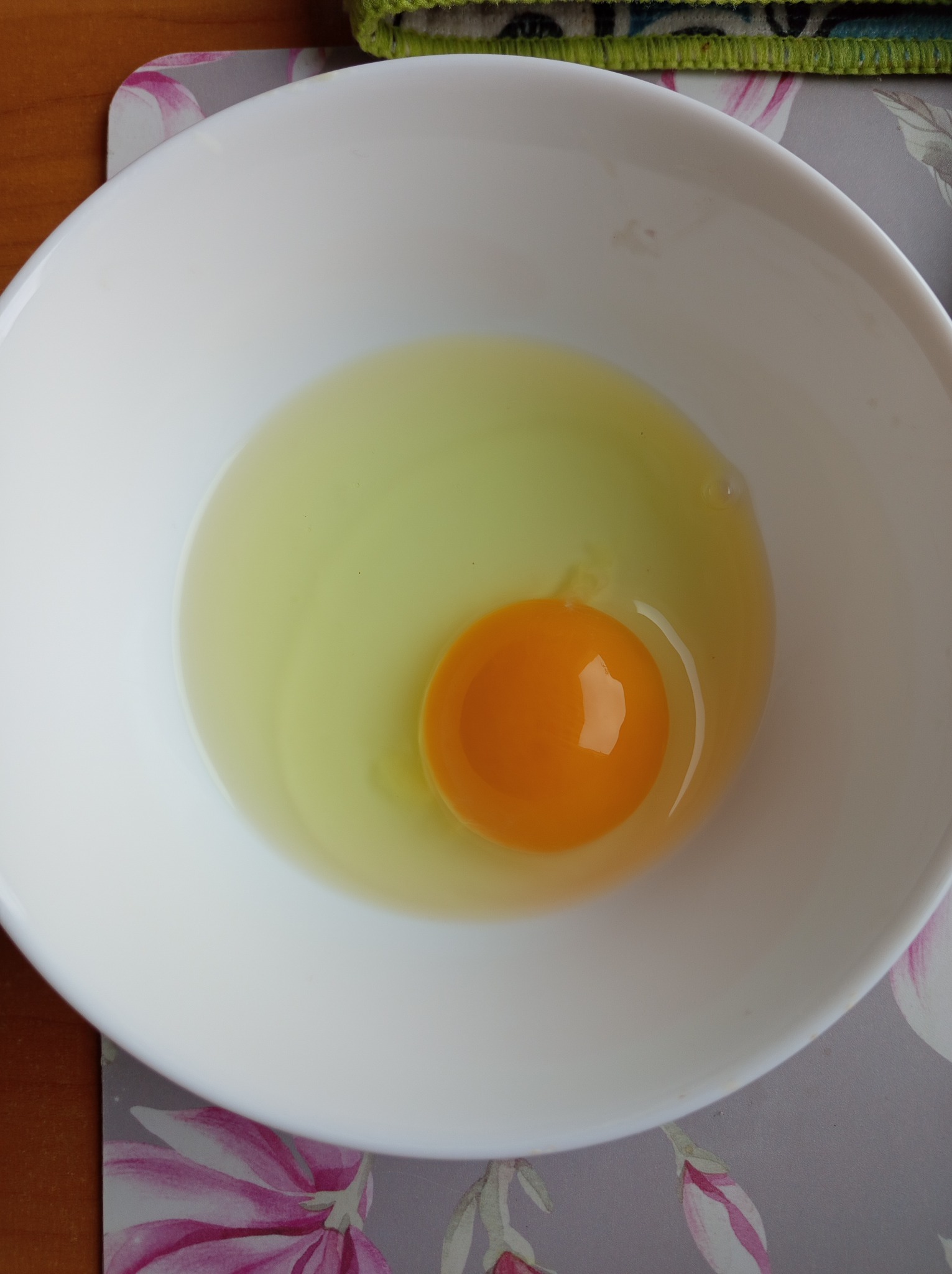 Surowe jajo z intensywnie pomarańczowym żółtkiem w białej miseczce na podkładce z motywem kwiatowym.