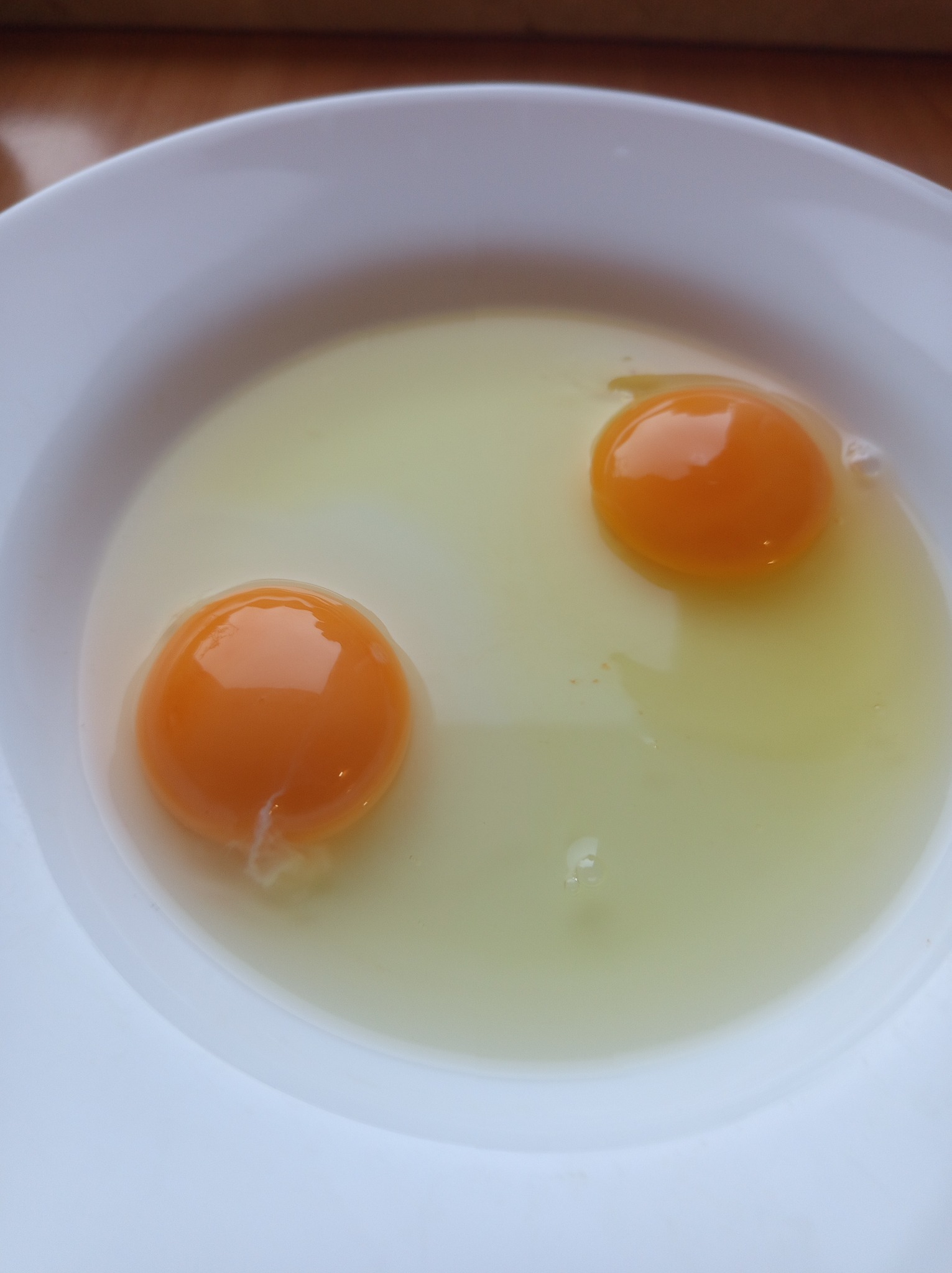 Dwa surowe żółtka z resztkami białka w białej misce, uchwycone z góry w neutralnym oświetleniu.