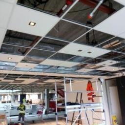Profesjonalne Usługi Budowlane - Montaż sufitu podwieszanego kasetonowego z widoczną konstrukcją metalową i instalacjami; pracownik na drabinie w odblaskowej kamizelce.