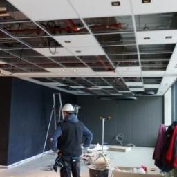 Profesjonalne Usługi Budowlane - Montaż sufitu podwieszanego w biurze, widoczne elementy konstrukcyjne i instalacje, pracownik w kasku na tle drabiny.