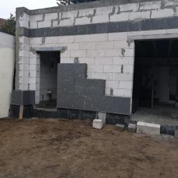 Profesjonalne Usługi Budowlane - Budynek w trakcie ocieplania, widoczne białe bloczki konstrukcyjne i szare płyty izolacyjne częściowo zamontowane na ścianie. Na pierwszym planie ziemia.