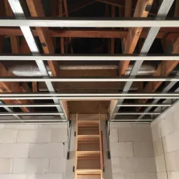 Montaż sufitu podwieszanego z widoczną konstrukcją metalową i drewnianymi belkami stropowymi oraz składanymi schodami strychowymi, na ścianie z białych bloczków.