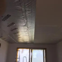 Częściowo ocieplony sufit z folią aluminiową marki SECCO, widoczne łączenia i fragment sufitu z płytami, dwie aluminiowe drabiny ustawione przy oknie.