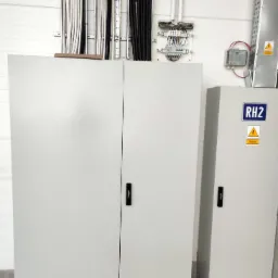 Dwie białe szafy elektryczne z czarnymi uchwytami w hali przemysłowej, widoczne kable w korytach na ścianie oraz schemat elektryczny z przewodami.