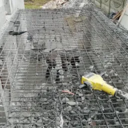 Demontaż zbrojenia stropu z gruzem, widoczny żółty młot pneumatyczny Bosch GSH 27, w tle drzewa i budynki.