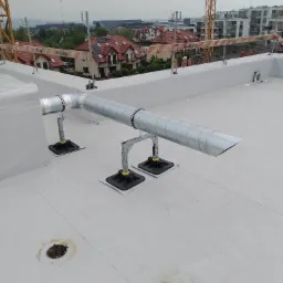 Srebrny kanał wentylacyjny spiralny na dachu budynku, wsparty na metalowych konstrukcjach z czarnymi podstawami, w tle zabudowa miejska i dźwig budowlany.