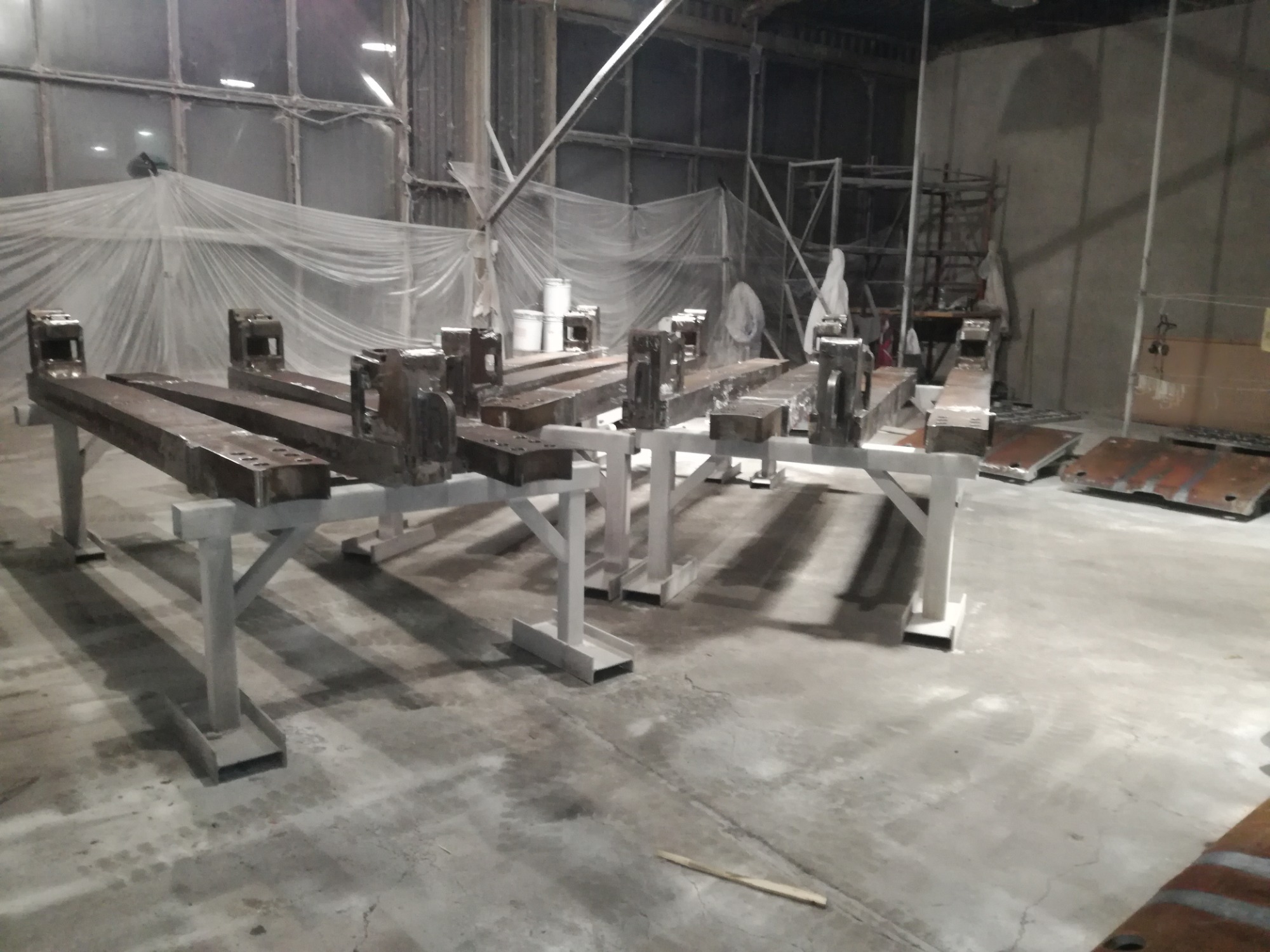 Metalowe konstrukcje na stojakach, po obróbce, prawdopodobnie przygotowane do dalszego etapu, widoczne ślady po piaskowaniu lub innym procesie czyszczenia, w tle hala produkcyjna z zabezpieczeniami.