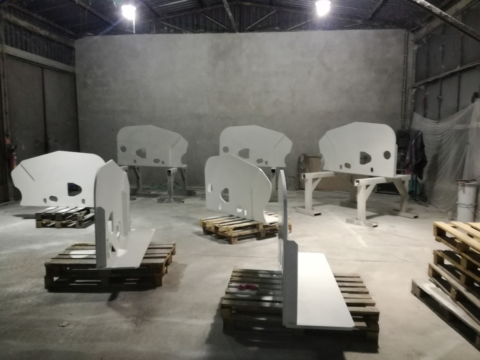 Białe, metalowe elementy konstrukcyjne z otworami, po malowaniu proszkowym, ułożone na paletach w hali produkcyjnej z surowymi ścianami.