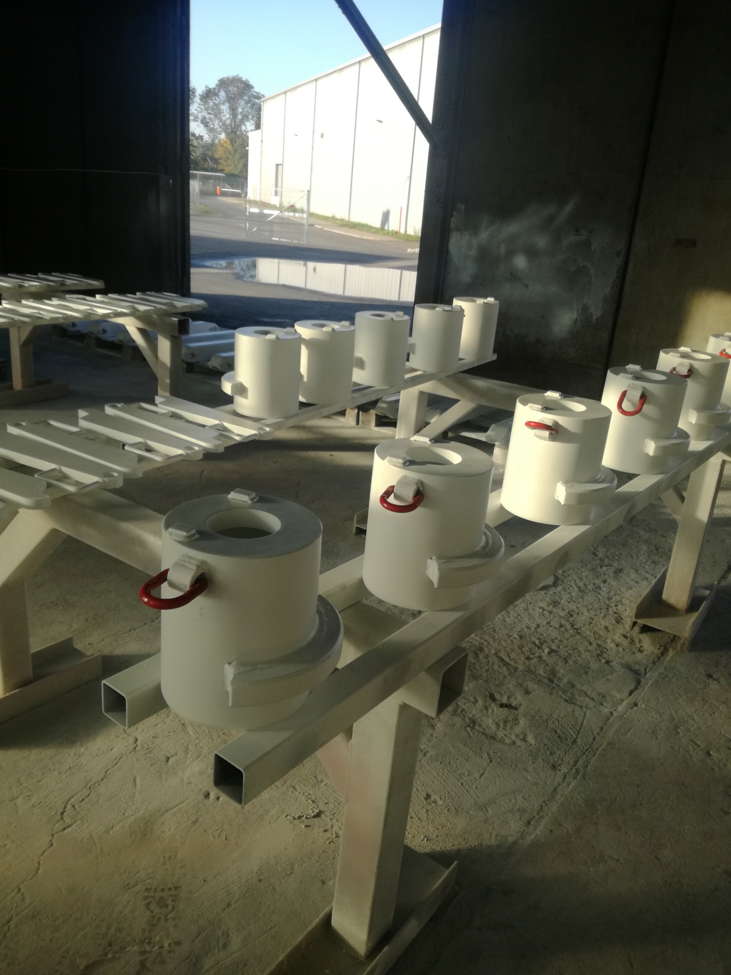 Rząd cylindrycznych, białych elementów z czerwonymi uchwytami, prawdopodobnie form odlewniczych, świeżo po malowaniu, ustawionych na metalowych stojakach w hali produkcyjnej.