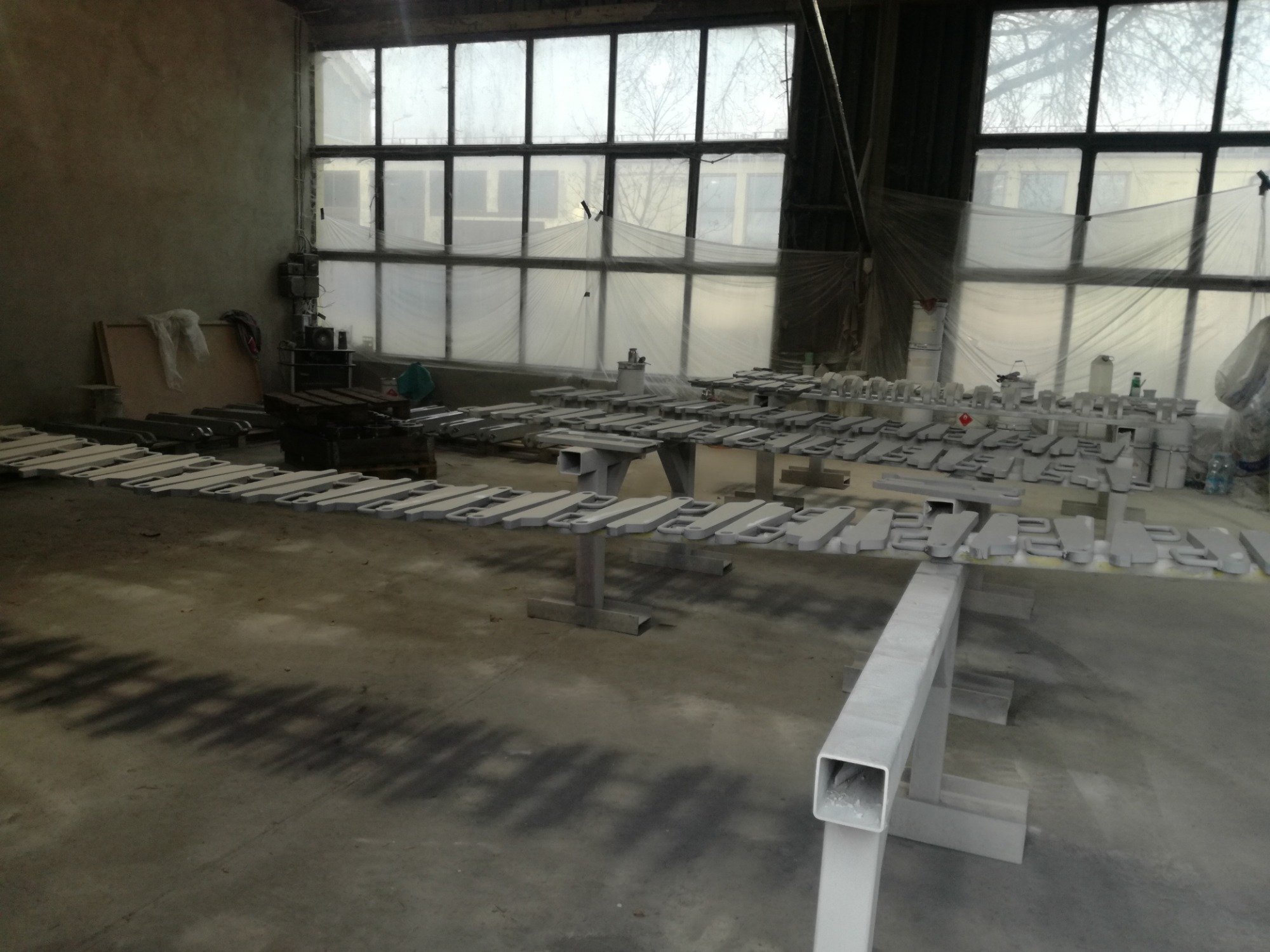 Metalowe elementy ułożone rzędami na stojakach po procesie obróbki powierzchni w hali produkcyjnej, widoczne ślady po malowaniu natryskowym, duże okna z widokiem na zewnątrz
