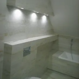 Wykończona łazienka w odcieniach beżu z podwieszaną toaletą, wanną i oświetleniem punktowym w zabudowie nad półką.