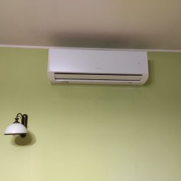 Mili Service - Biały klimatyzator zamontowany na oliwkowozielonej ścianie, obok kinkiet w stylu retro z białym kloszem.