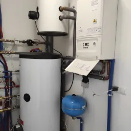 Biały, wiszący kocioł gazowy z otwartą klapą serwisową, obok biały zasobnik ciepłej wody z czarnym osprzętem, niebieskie naczynie przeponowe i liczne rury miedziane oraz izolowane na niebiesko...