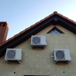Trzy klimatyzatory zamontowane na elewacji domu jednorodzinnego, widoczne ślady po wcześniejszych instalacjach, ceglany komin i tr&oacute;jkątne okno na poddaszu.