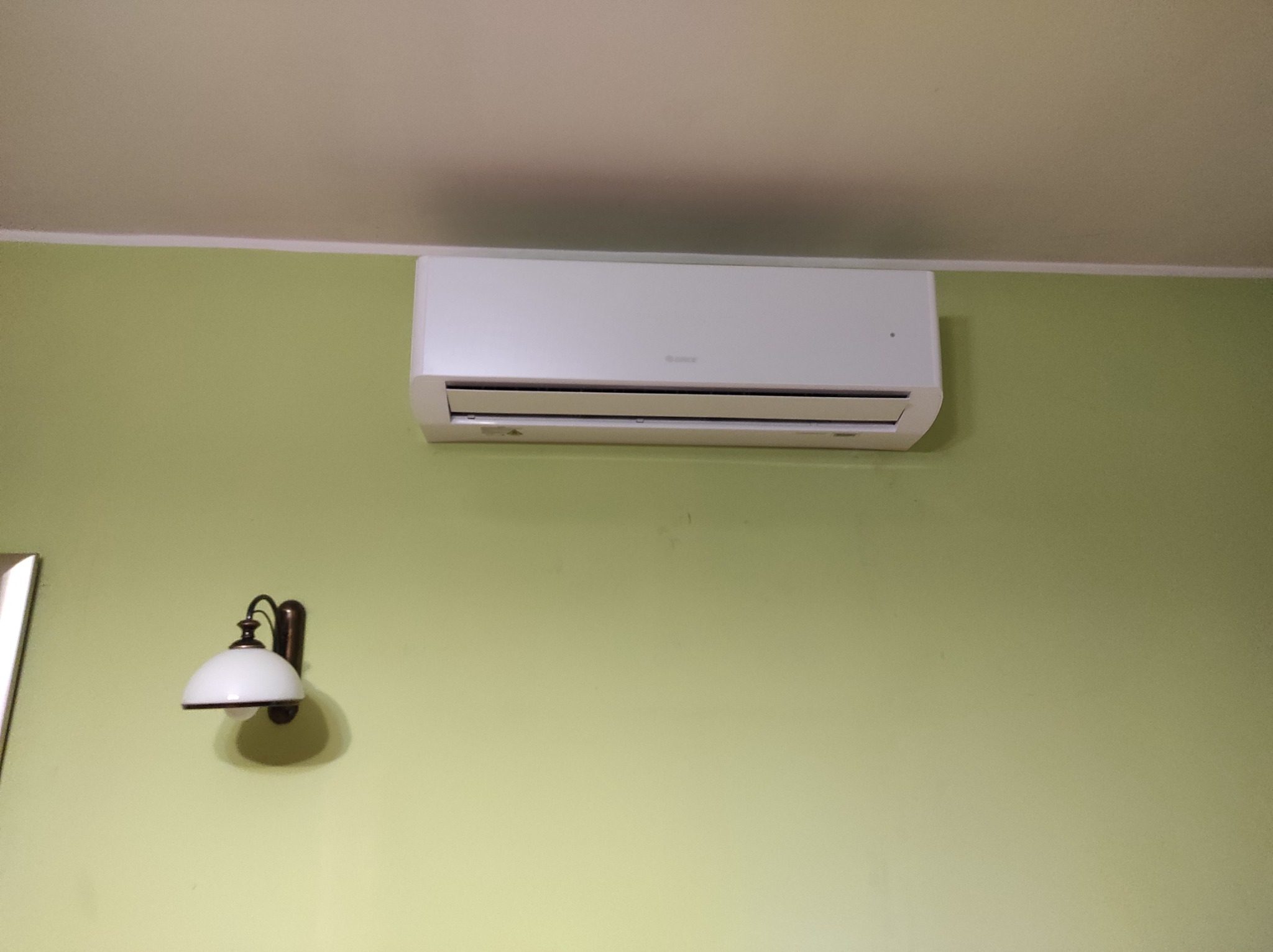Biały klimatyzator zamontowany na oliwkowozielonej ścianie, obok kinkiet w stylu retro z białym kloszem.