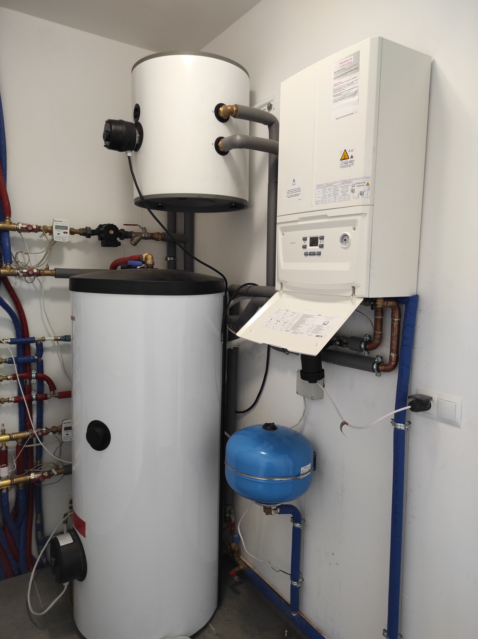Biały, wiszący kocioł gazowy z otwartą klapą serwisową, obok biały zasobnik ciepłej wody z czarnym osprzętem, niebieskie naczynie przeponowe i liczne rury miedziane oraz izolowane na niebiesko...