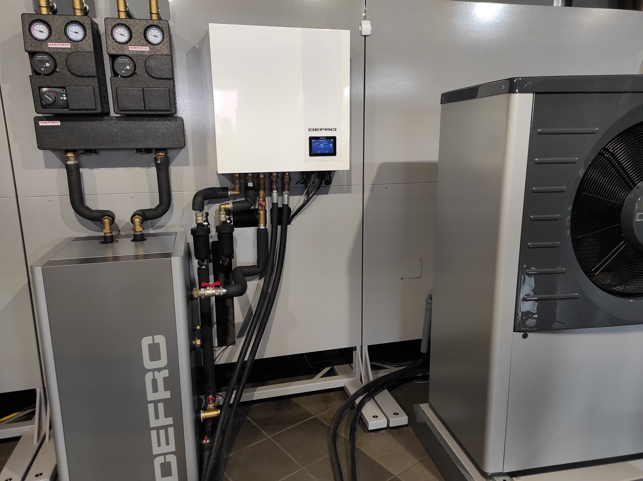 Kompletna instalacja pompy ciepła DEFRO z widocznymi elementami: jednostką wewnętrzną z wyświetlaczem, modułem hydraulicznym, czujnikami ciśnienia oraz jednostką zewnętrzną z wentylatorem.