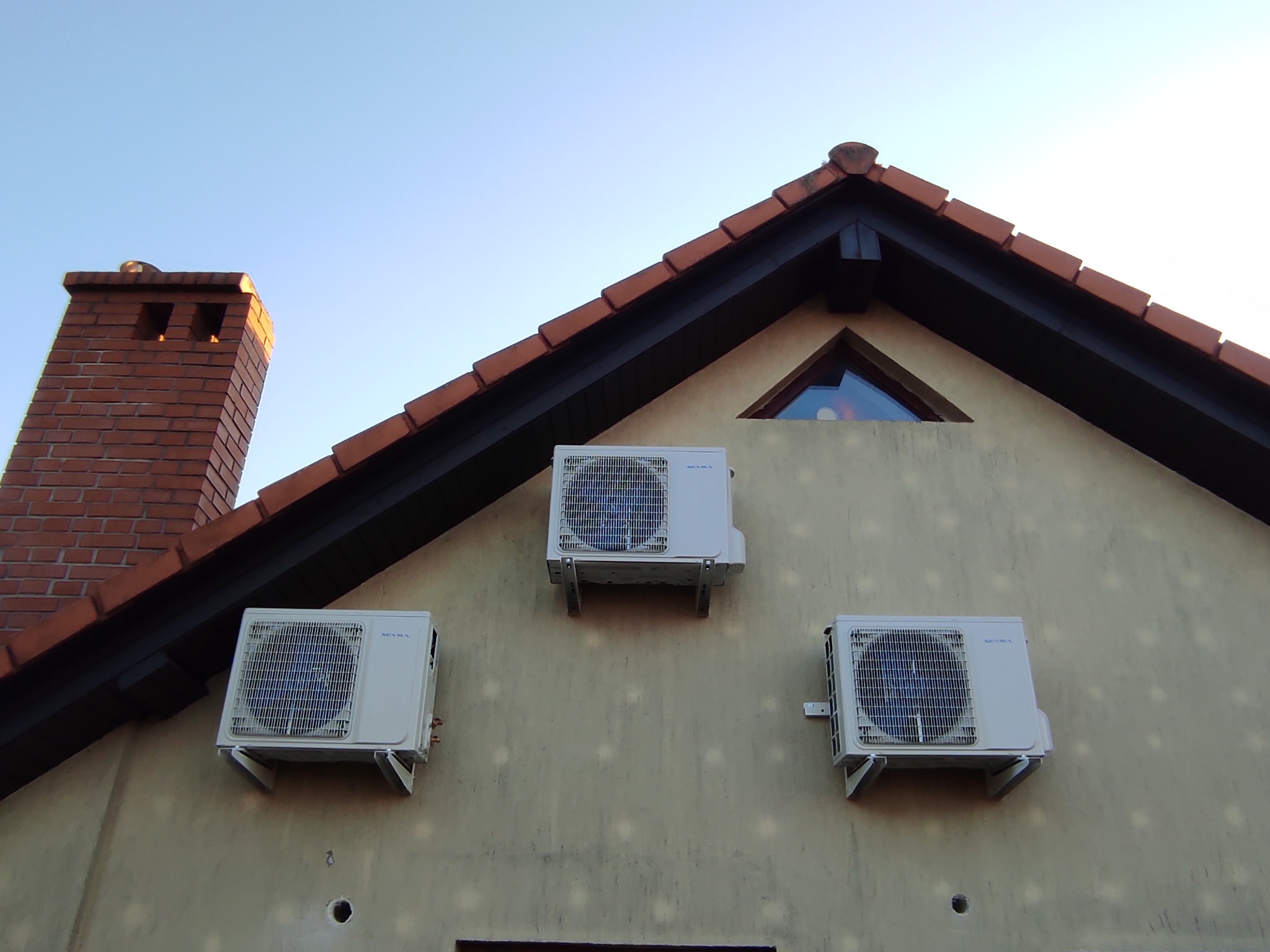 Trzy klimatyzatory zamontowane na elewacji domu jednorodzinnego, widoczne ślady po wcześniejszych instalacjach, ceglany komin i trójkątne okno na poddaszu.
