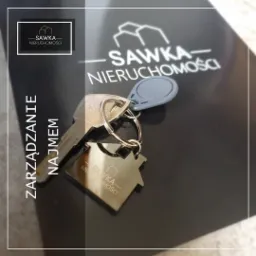 Klucz do mieszkania z brelokiem w kształcie domu z logo agencji nieruchomości Sawka, leżący na czarnym folderze z nadrukowanym logo firmy. Widoczny napis 'Zarządzanie najmem'.