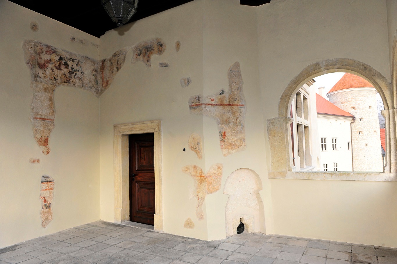 Wnętrze zamku w Pieskowej Skale przed konserwacją polichromii XVIII i XVI wieku, widoczne fragmenty malowideł na ścianach, kamienna podłoga i okno z widokiem na dziedziniec.