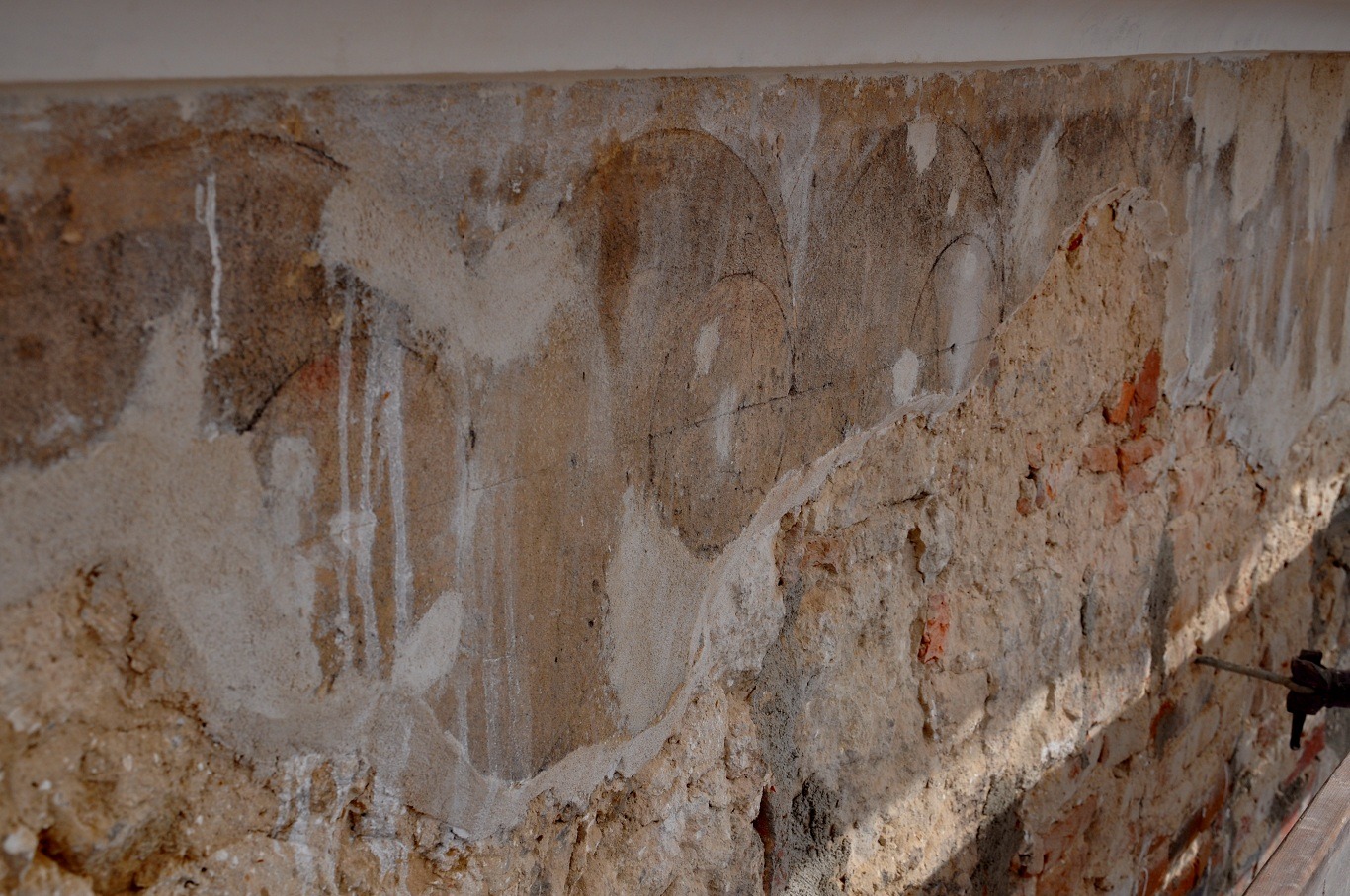 Fragment ściany z odsłoniętymi warstwami tynku i cegły, widoczne ślady polichromii w postaci zarysowanych łuków, ślady prac konserwatorskich.