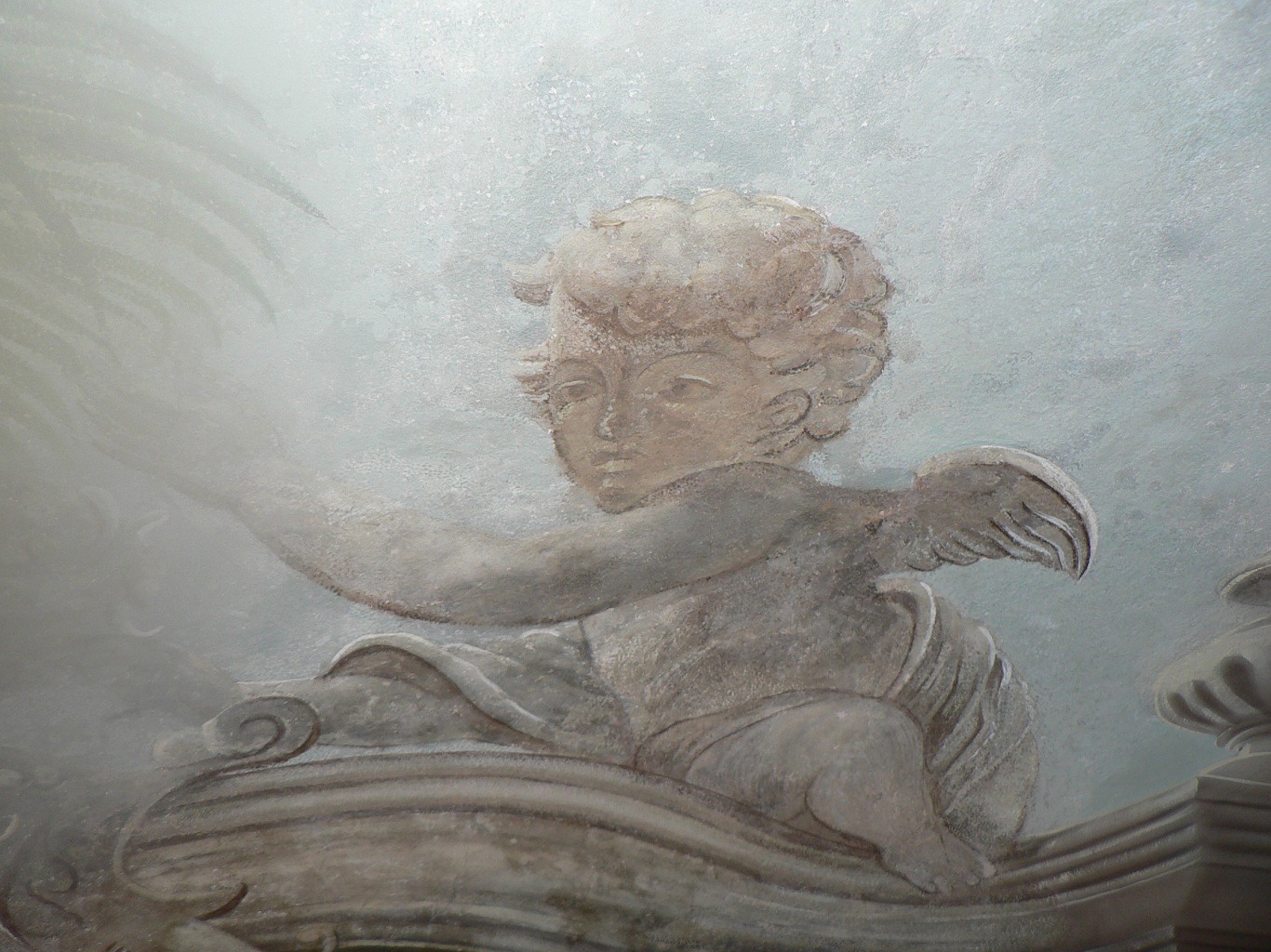 Detal malowidła sufitowego przedstawiającego aniołka z rozwianymi włosami i drobnymi piórami na skrzydłach, siedzącego na ozdobnym elemencie architektonicznym, w odcieniach beżu i szarości.