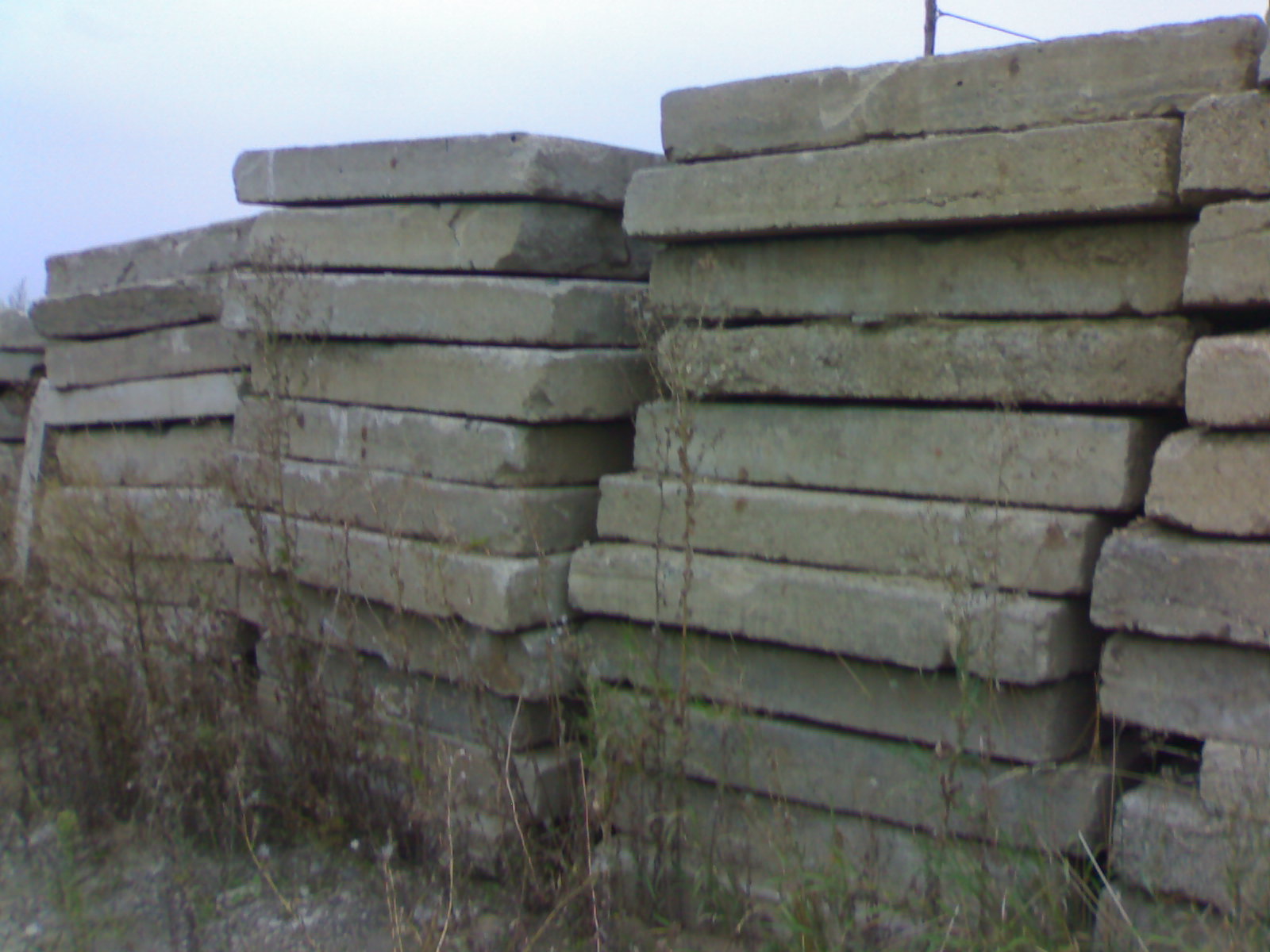 Stosy szarych, betonowych płyt drogowych ułożonych na zewnątrz, z widocznymi drobnymi roślinami wyrastającymi spomiędzy nich, sfotografowane w pochmurny dzień.