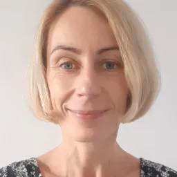 Portret kobiety z krótkimi blond włosami, łagodny uśmiech, na jasnym tle.