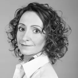 Portret kobiety z kręconymi włosami, ubranej w białą koszulę, z delikatnym uśmiechem i patrzącej w obiektyw, czarno-białe zdjęcie.