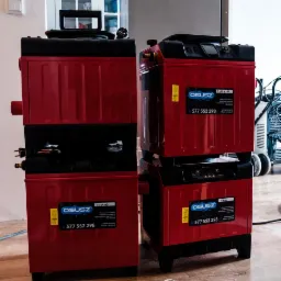 Dwa czerwone, modułowe urządzenia do osuszania podłogowego stoją na drewnianej podłodze w pomieszczeniu. Na każdym urządzeniu naklejka z logo i numerem telefonu.