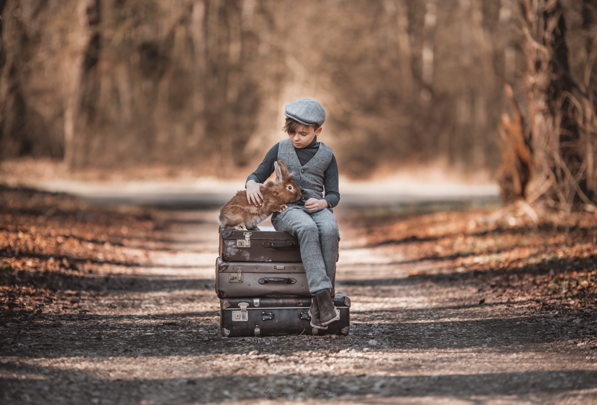 Chłopiec w tweedowym garniturze i kaszkiecie siedzi na stosie starych walizek, głaszcząc brązowego królika na leśnej ścieżce.